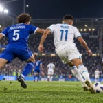 Joueurs de Troyes et de Marseille vus de dos pendant une action au stade, avec les noms Maronnier et Harit – reconstitution générée par IA, pas une photo du match.